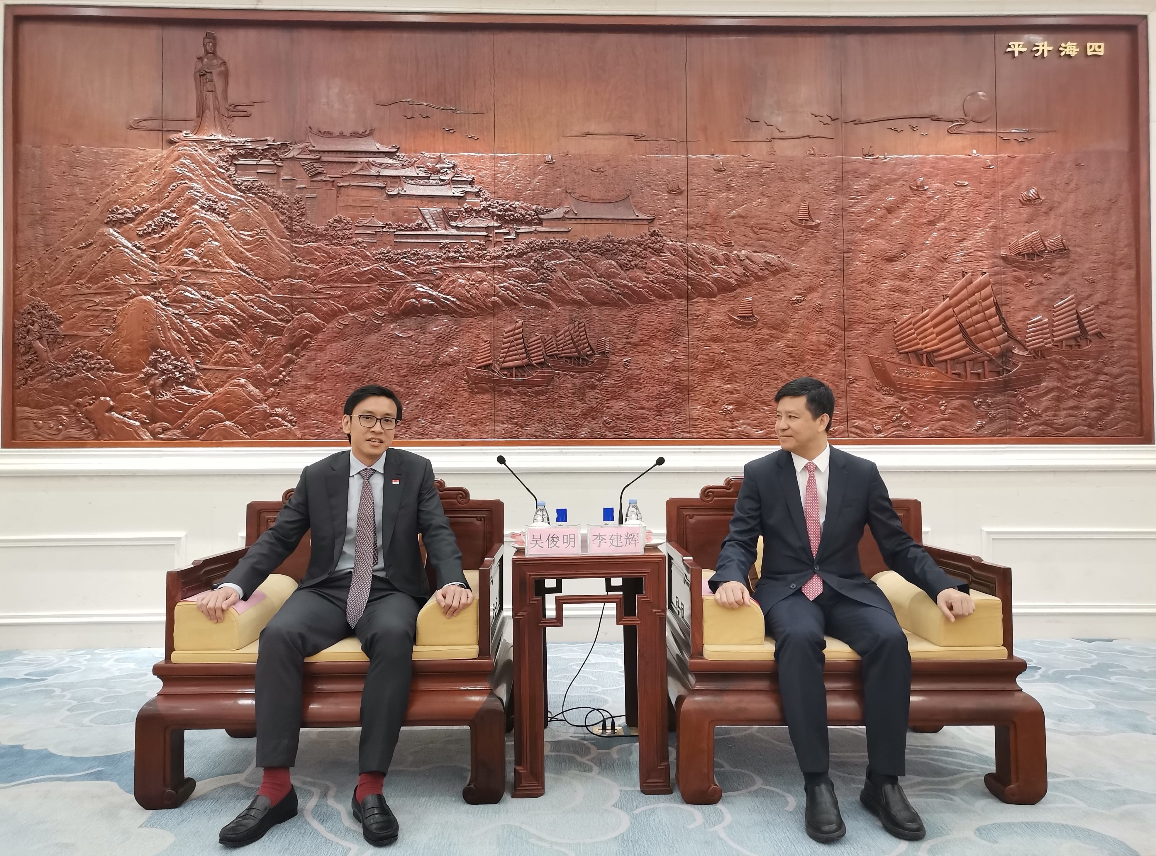 Two men in suits sit in ornate chairs before a carved wood wall panel depicting boats and landscape.